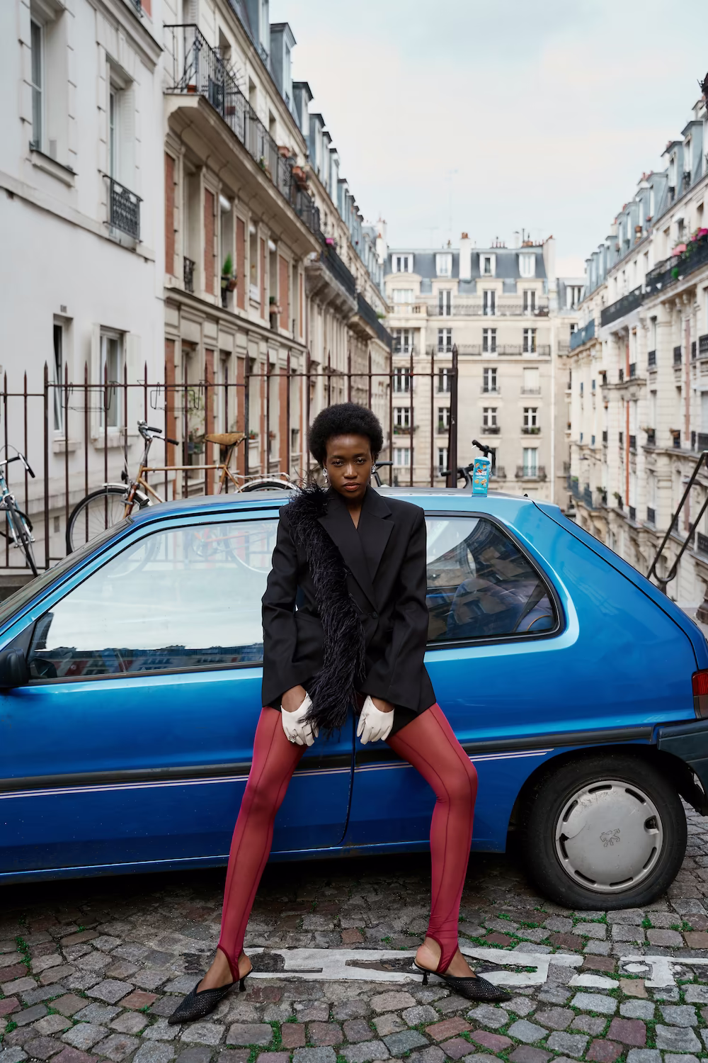 Model in an oversized black blazer, white gloves and red tights beside a blue vintage car in Paris — fashion editorial