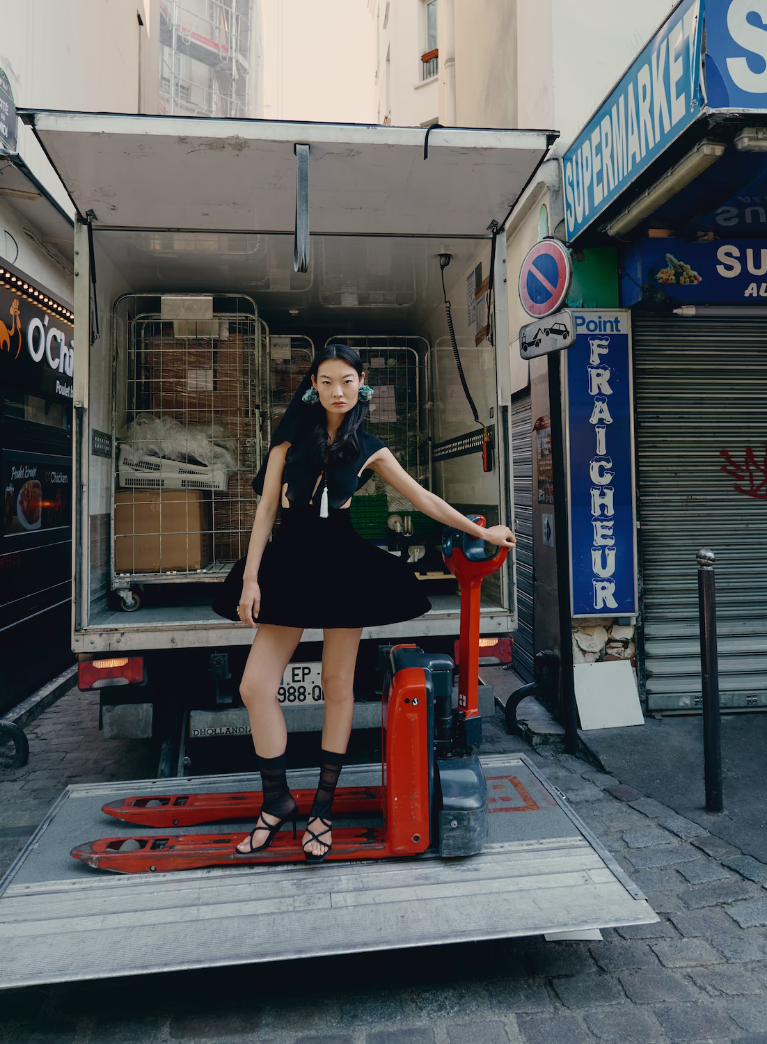 Model in a black dress standing on a delivery truck tail-lift outside a Paris supermarket — fashion editorial