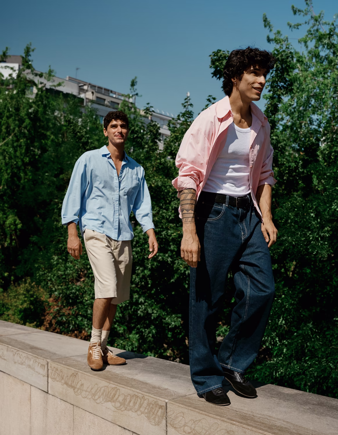 Two male models walking along a low wall in a pink shirt and a blue shirt, greenery behind — menswear lookbook
