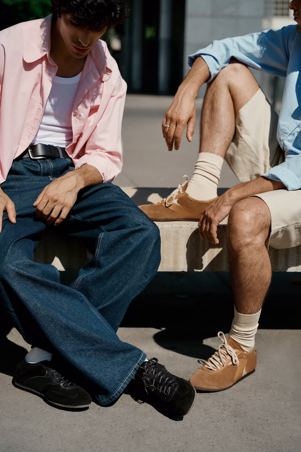 Two male models in a pink shirt and blue shirt seated on a stone bench in tan suede sneakers — menswear lookbook