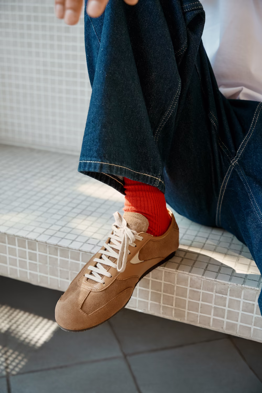 Close-up of wide dark denim over red socks and a tan suede sneaker on a tiled step — footwear product detail
