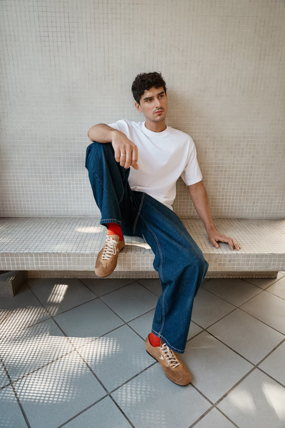 Male model in a white tee and wide jeans seated on a tiled bench in tan sneakers — menswear lookbook