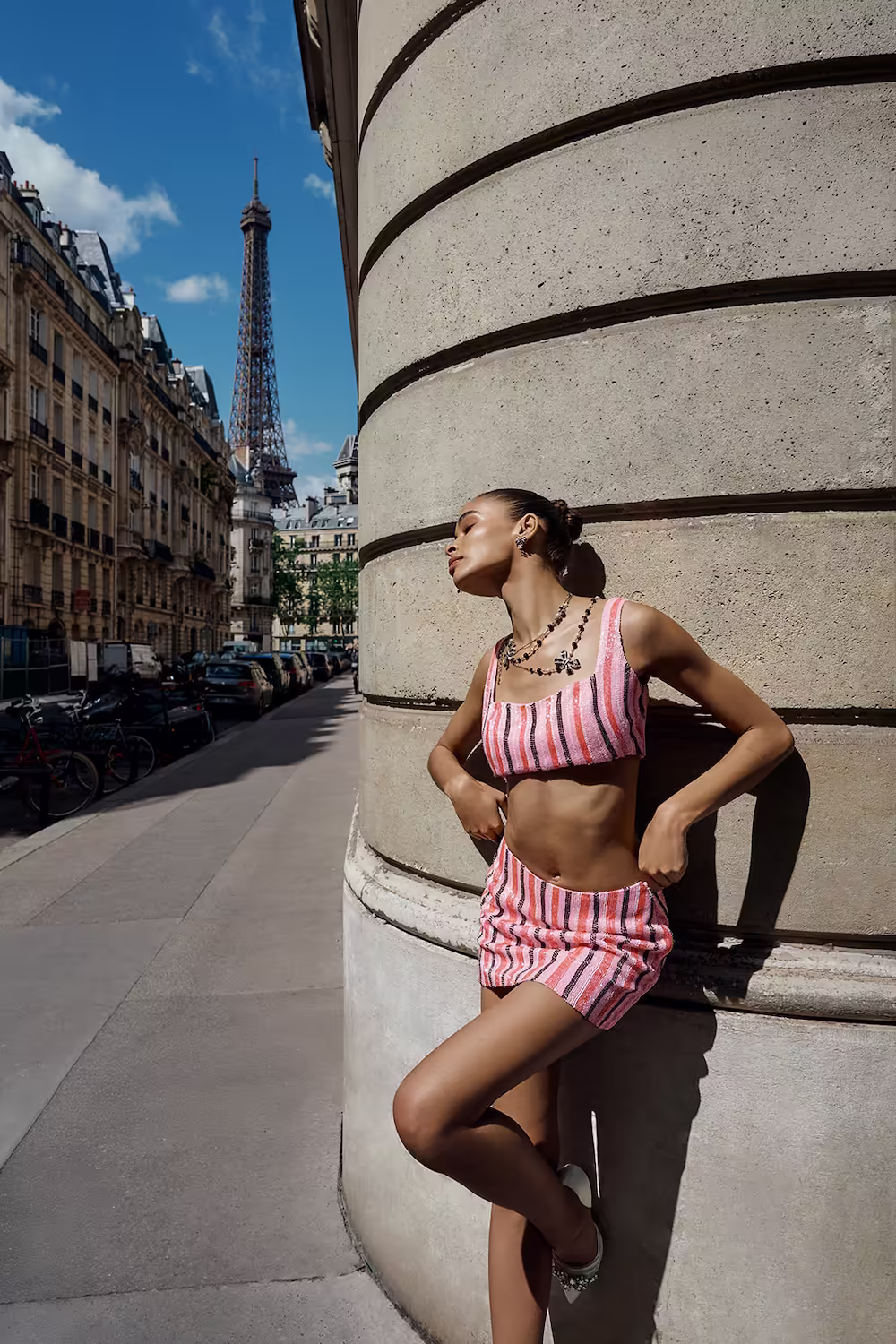 Model in a pink striped bra top and mini skirt against a fluted column, Eiffel Tower behind — fashion campaign