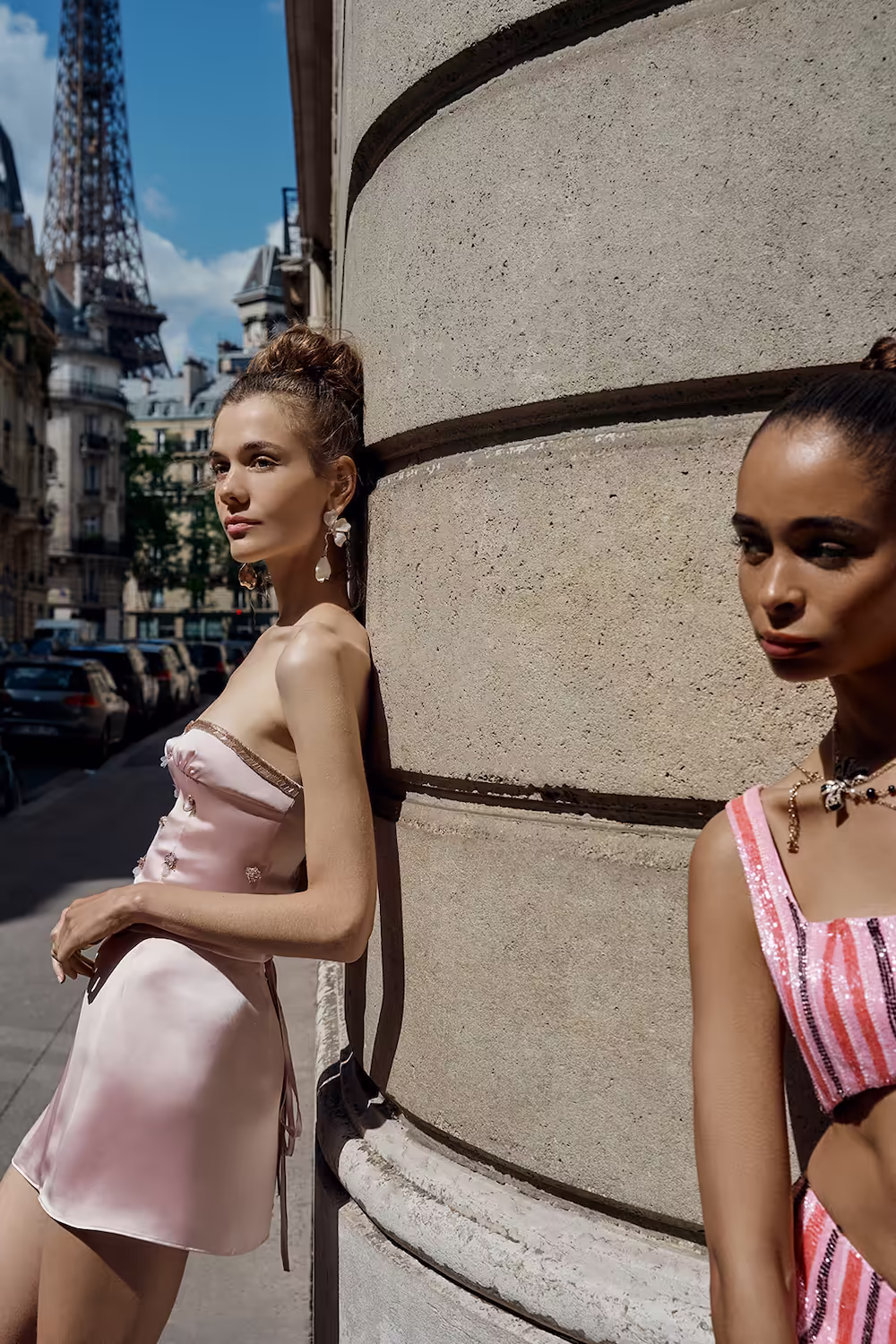 Two models in a pink satin slip dress and a striped bra top by a stone column, Eiffel Tower behind — fashion campaign