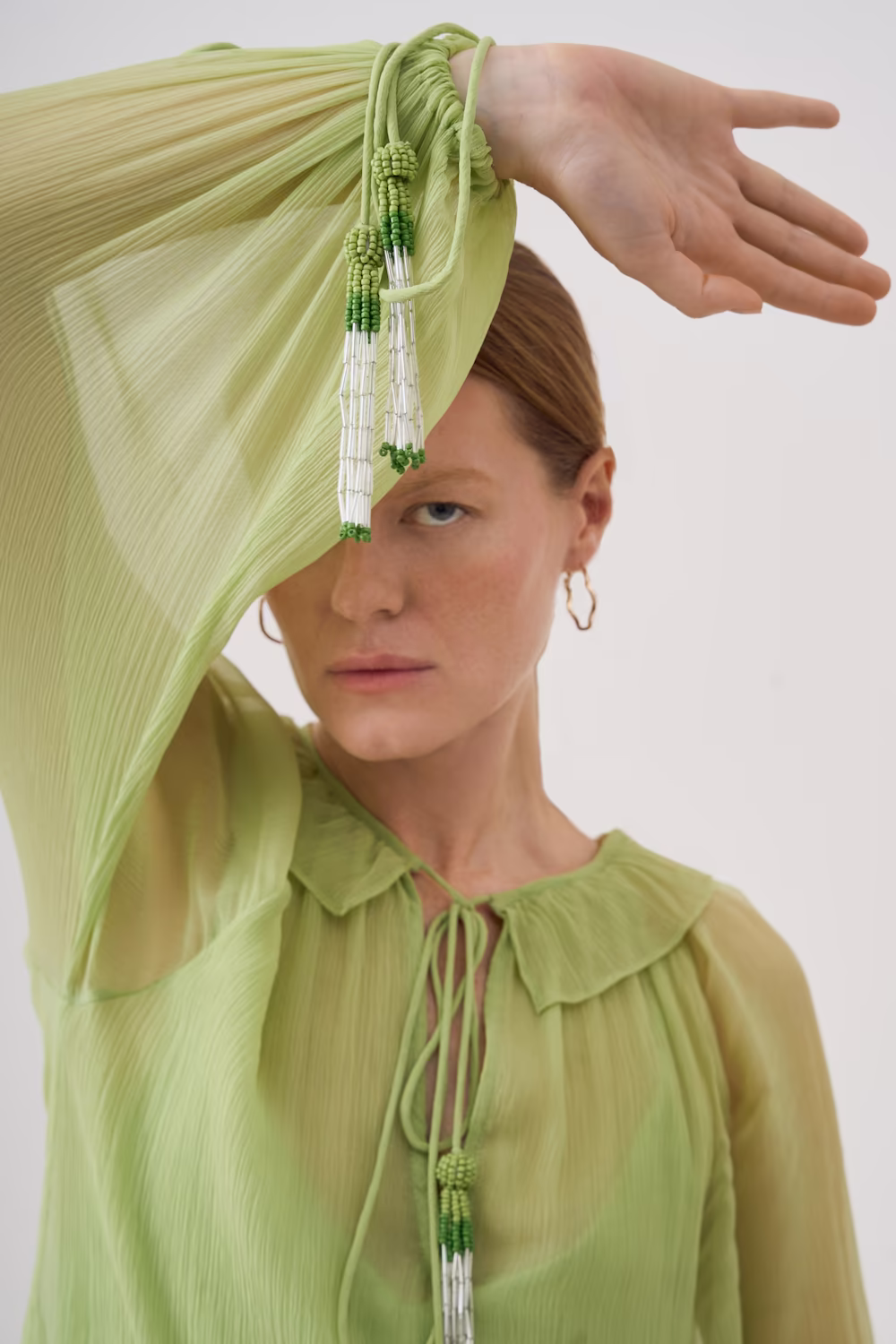 Model draping a lime-green pleated chiffon dress over her face, green tie-collar blouse beneath — fashion campaign