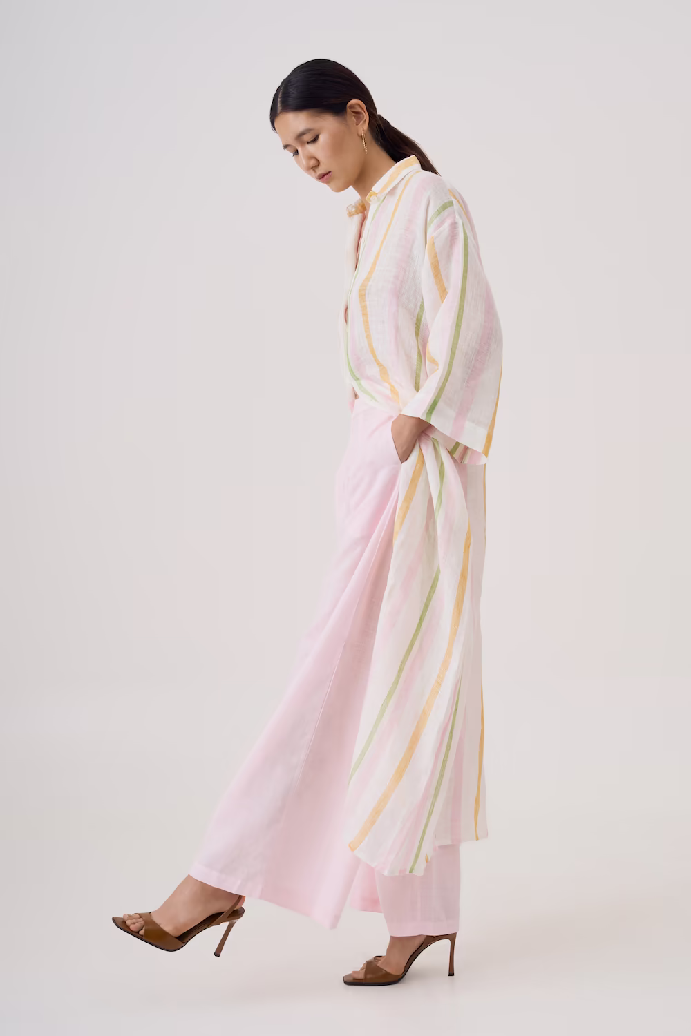 Model in a long pale pink and yellow striped sheer dress with brown heels on a plain backdrop — lookbook