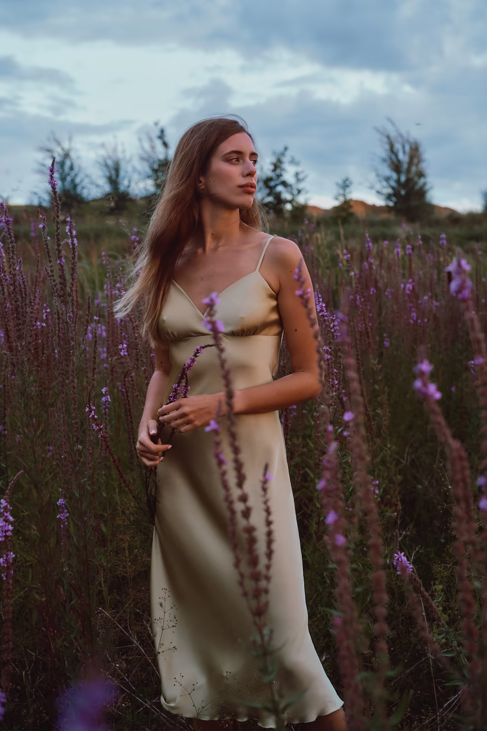 Model in a champagne satin slip dress standing among tall purple loosestrife — lifestyle fashion shoot