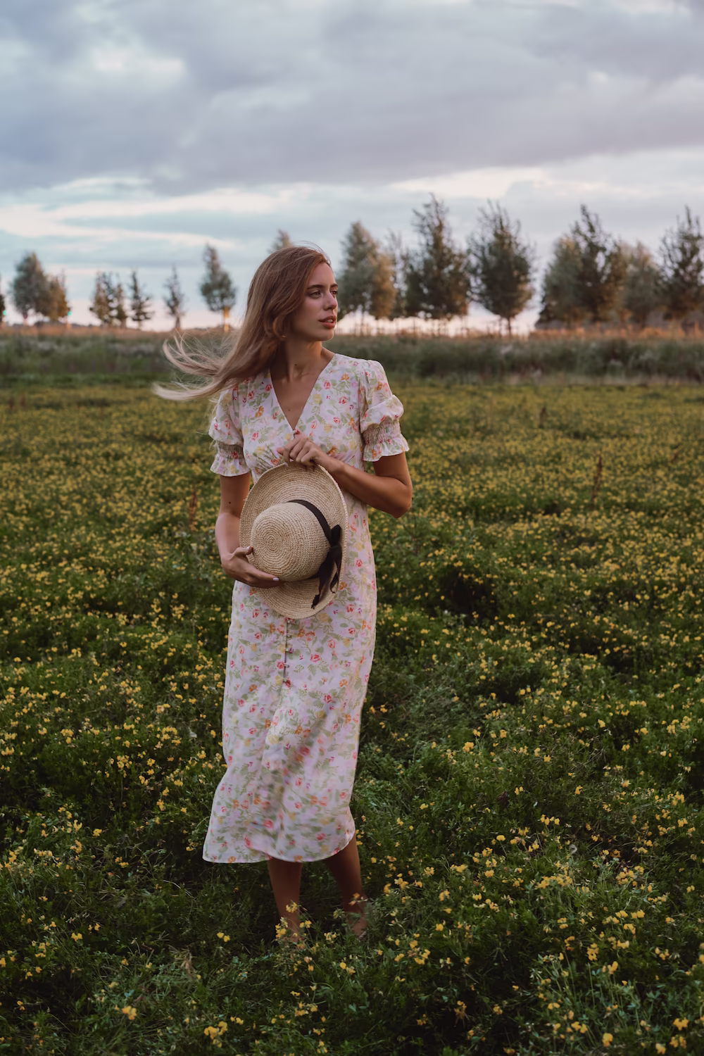 Model in a pink floral midi dress holding a straw hat in a flowering field at dusk — lifestyle campaign
