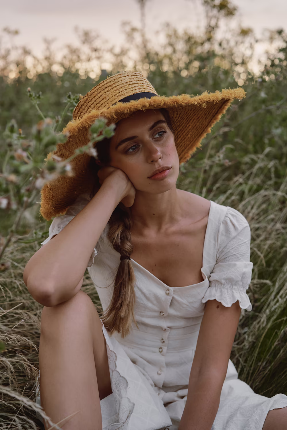 Model in a straw sun hat and white button dress seated in tall grass — lifestyle fashion shoot