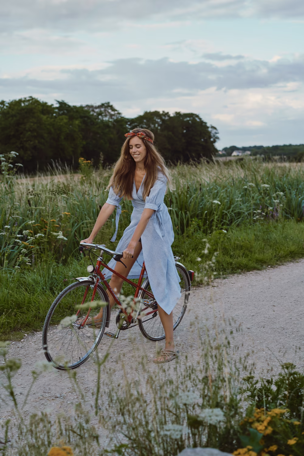 Model in a pale blue wrap dress riding a vintage bicycle on a gravel path through a meadow — lifestyle campaign