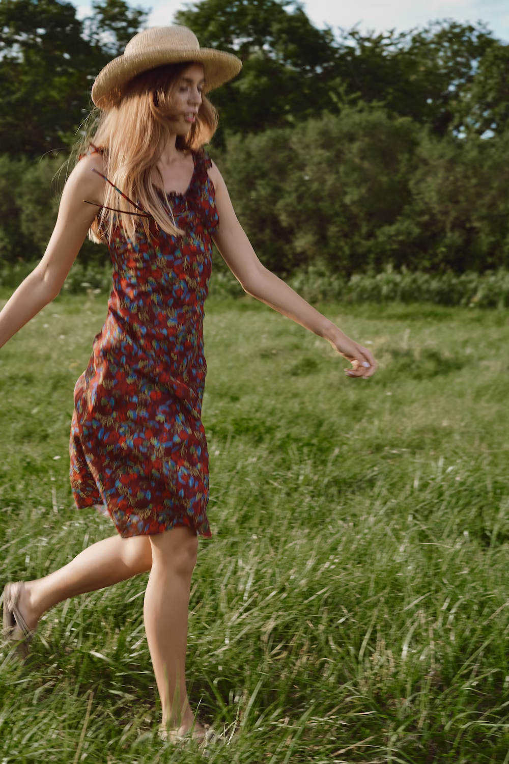 Model in a dark red floral mini dress and straw hat walking barefoot through summer grass — lifestyle campaign