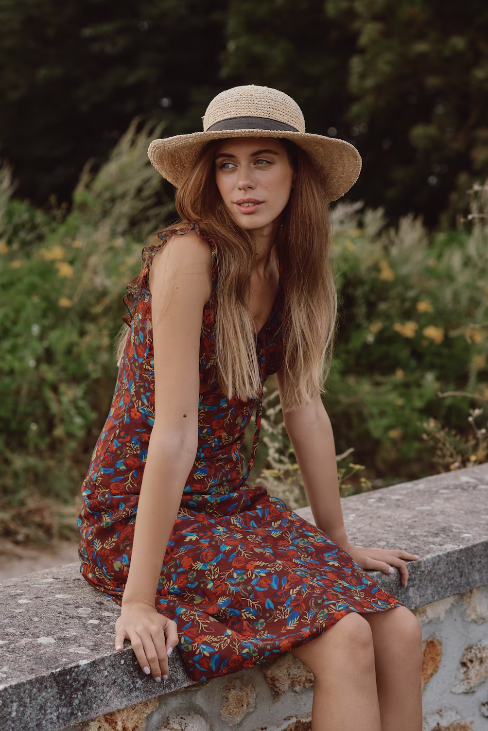 Model in a straw hat and red floral dress seated on a stone wall beside a meadow — lifestyle fashion shoot