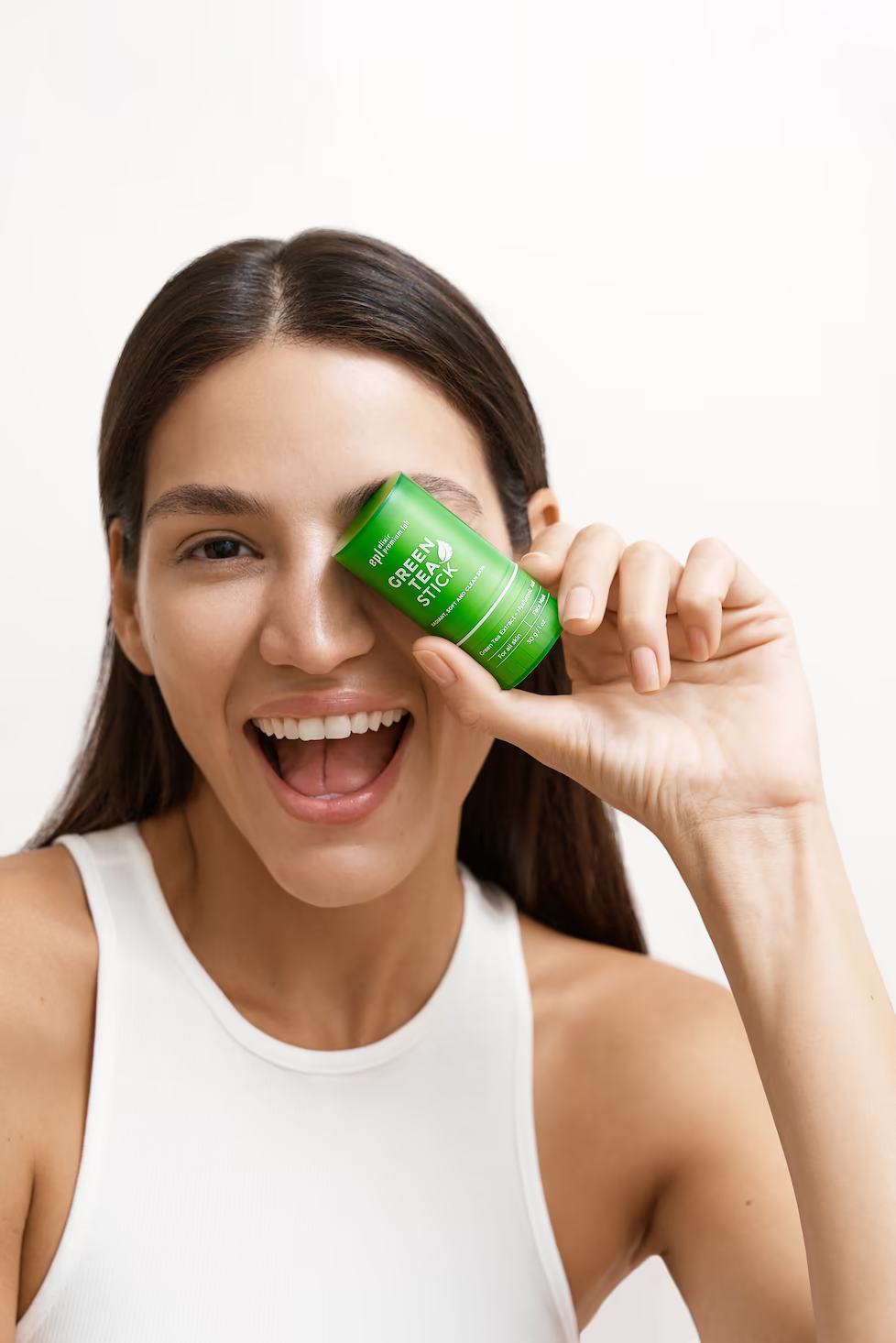 Beauty close-up of a laughing model in a white tank holding a green skincare stick over one eye — beauty campaign