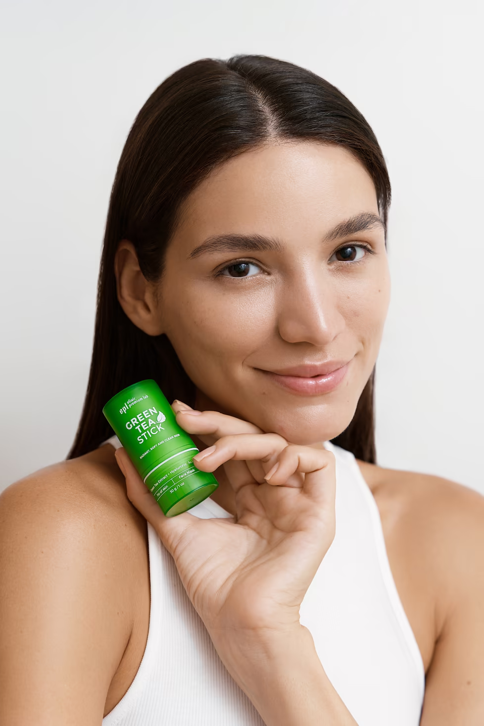 Beauty close-up of a smiling model in a white tank holding a green skincare stick at her neck — beauty campaign