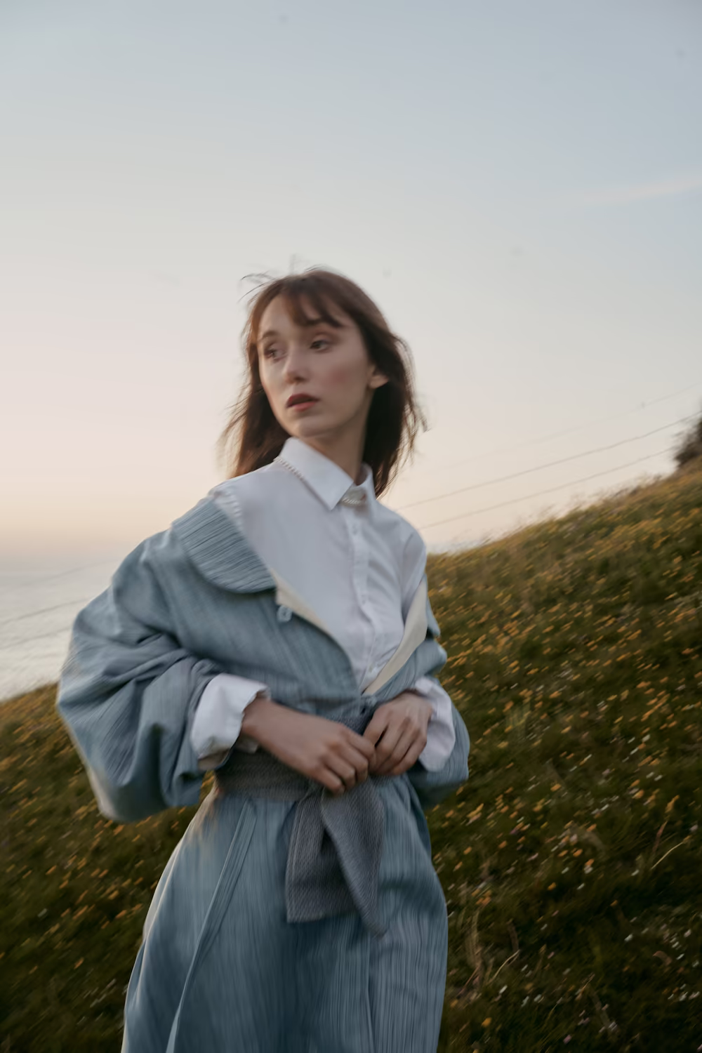 Model in a dusty-blue crinkled jacket over a white shirt in a coastal field at dusk — fashion lookbook