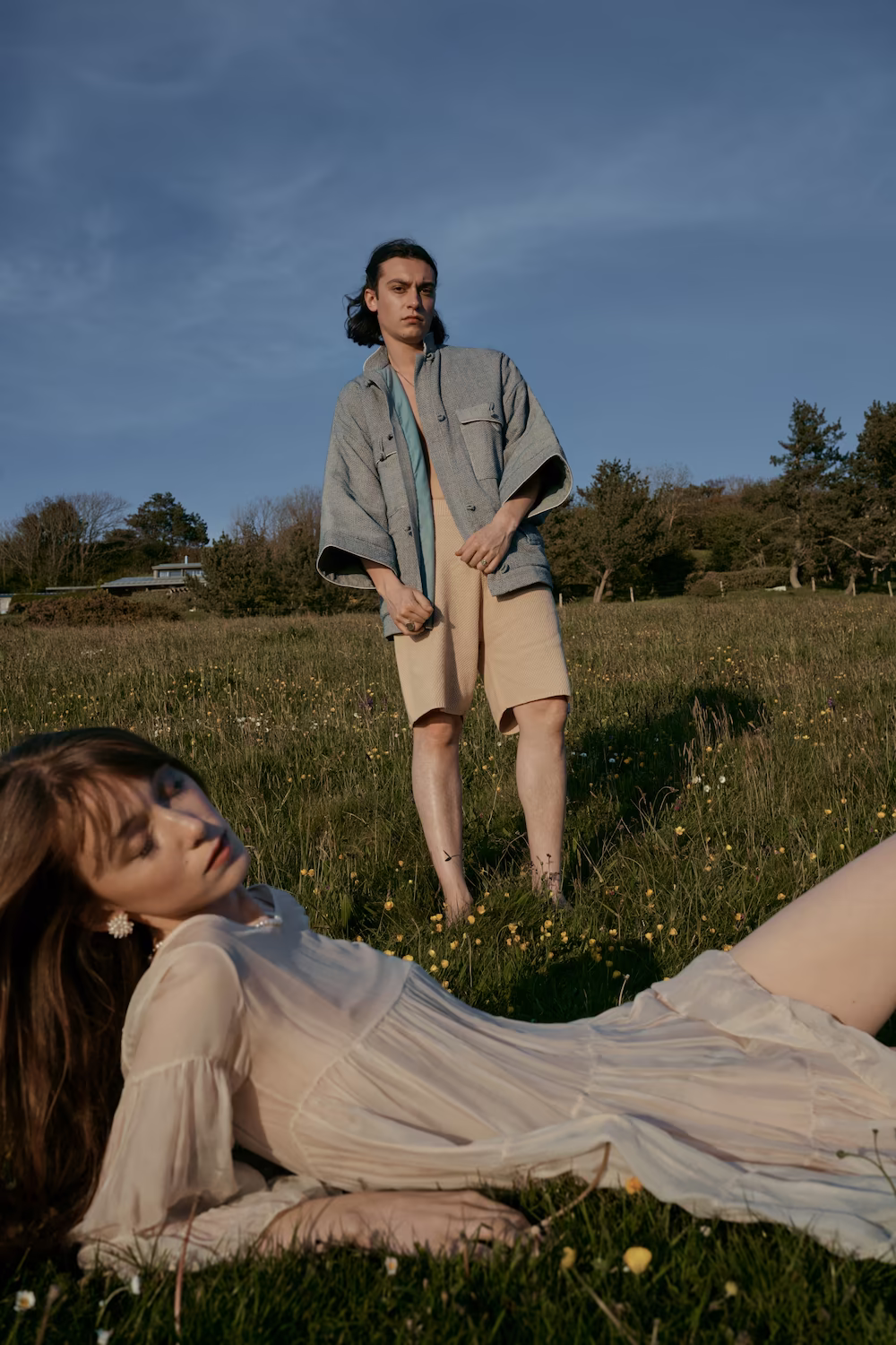 Male model standing in a meadow while a second model in a sheer cream dress reclines in the grass — fashion lookbook