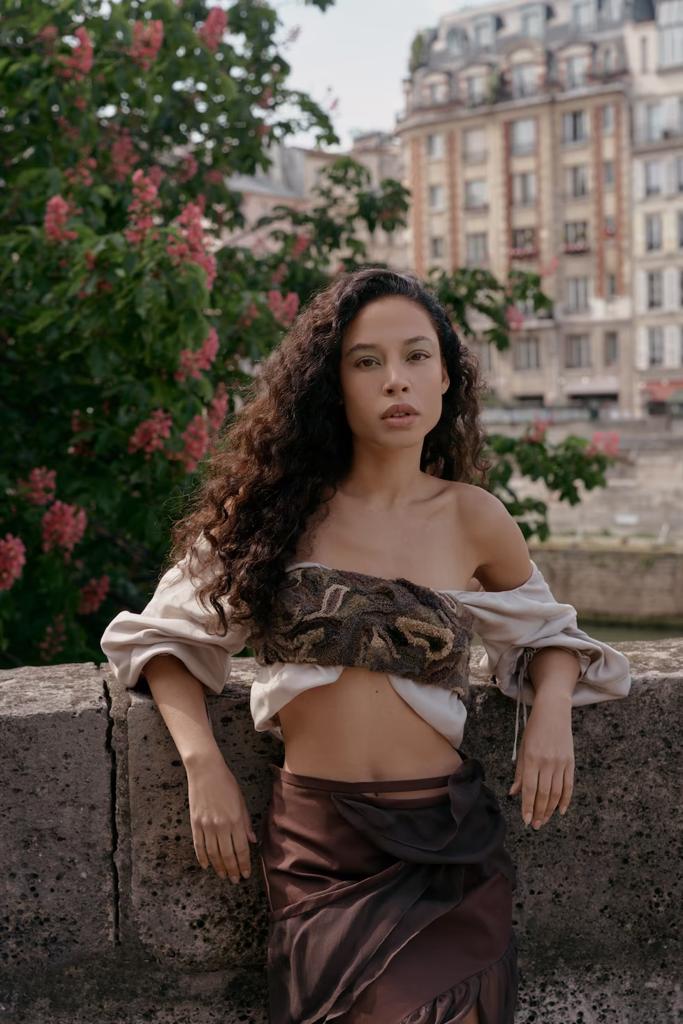 Model in an off-shoulder draped top and brown satin skirt on a Paris ledge with blossom — fashion campaign