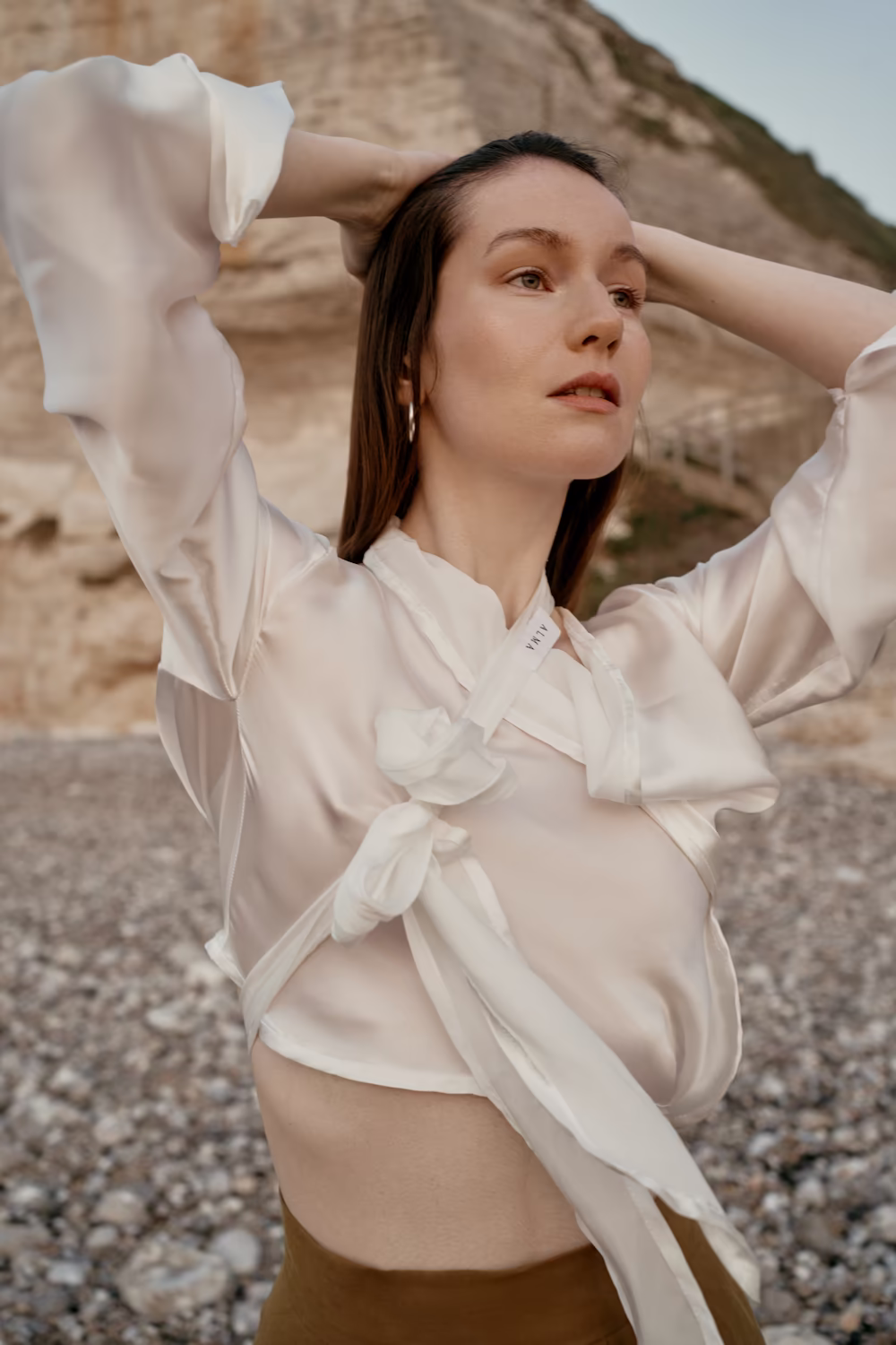 Model in a cream silk tie blouse with arms raised on a pebble beach below chalk cliffs — lifestyle campaign