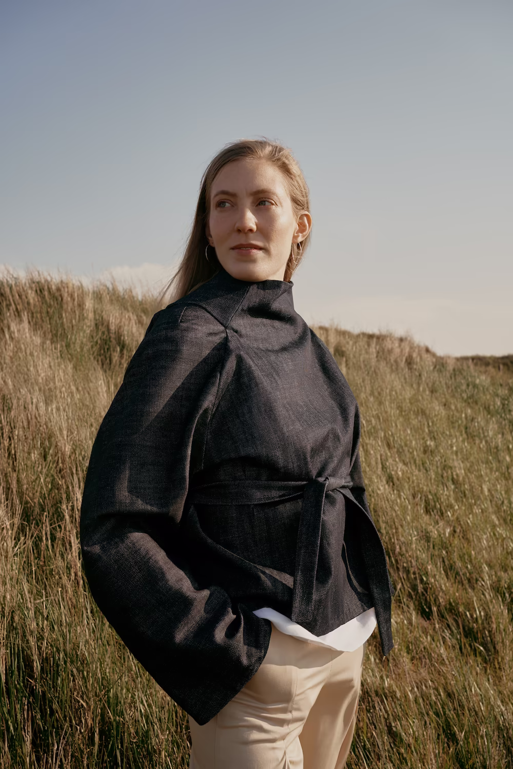 Model in an oversized black linen jacket and cream trousers standing in tall dune grass — fashion campaign