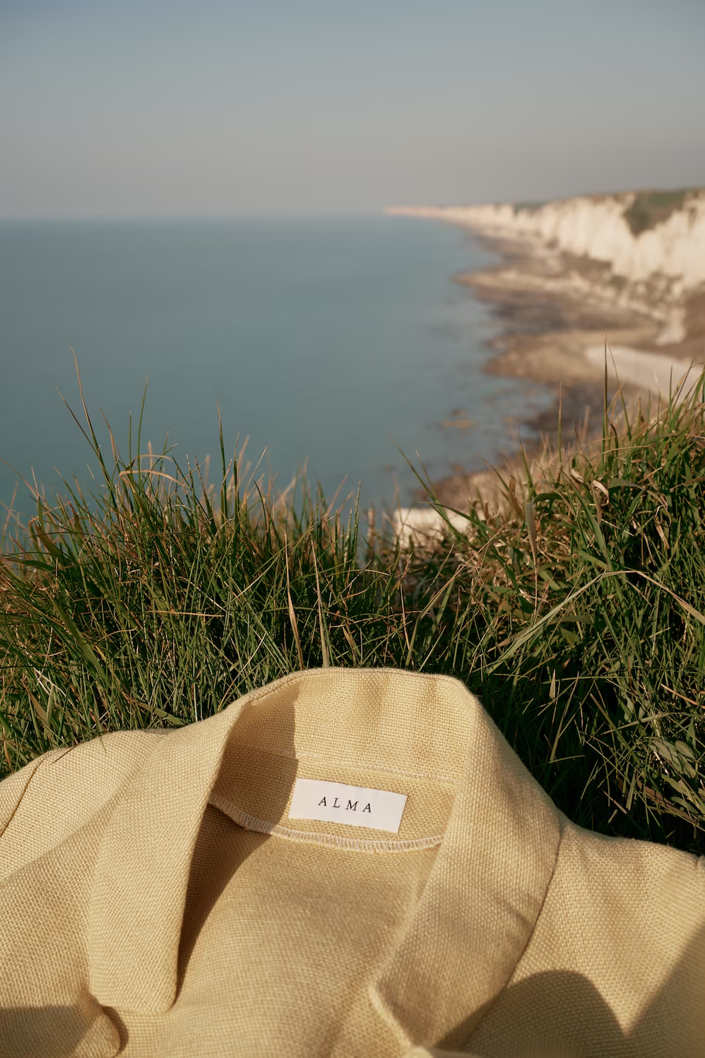 Folded pale yellow linen jacket laid on grass above chalk cliffs and the sea — product still life