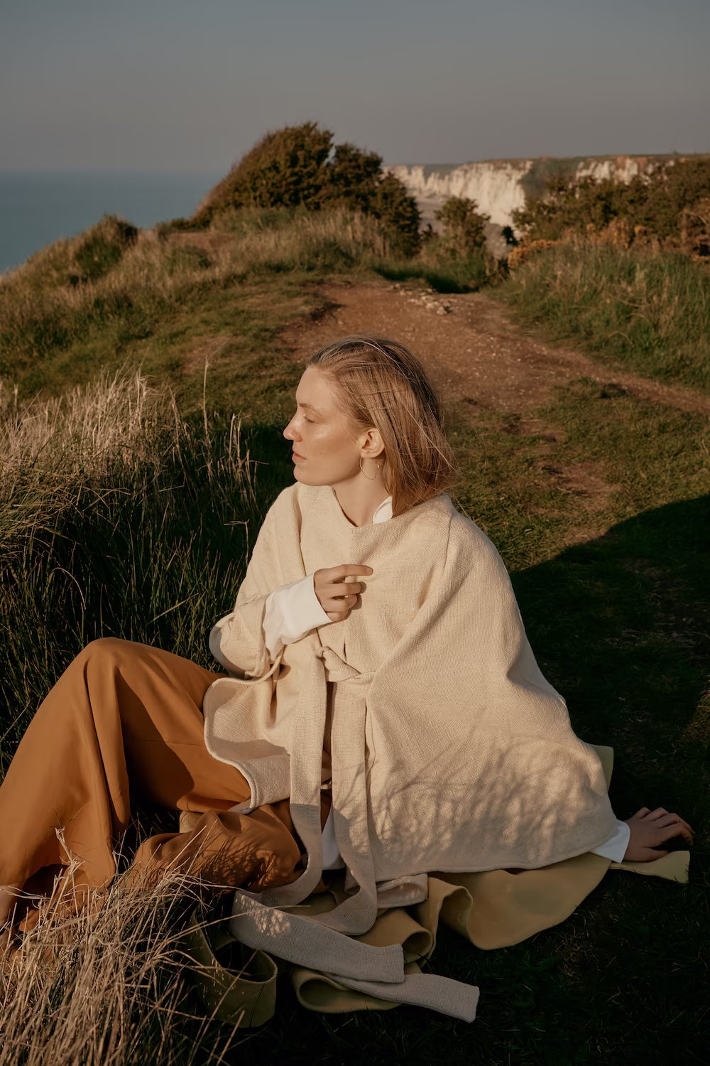 Model in a cream coat and camel trousers seated on a grassy clifftop path — lifestyle fashion campaign