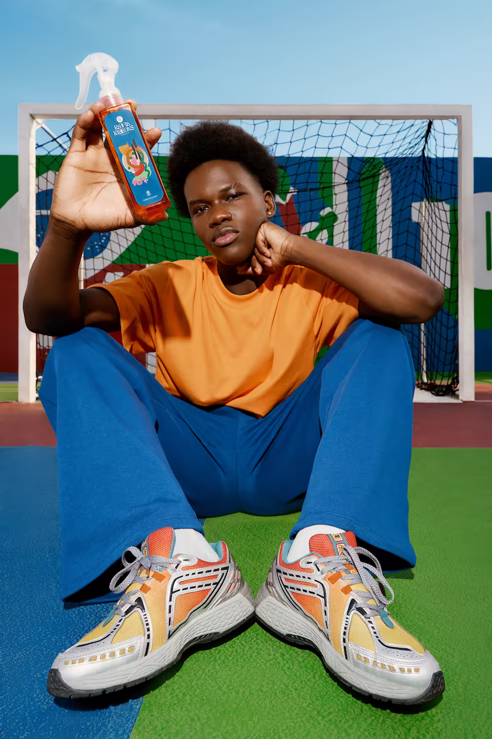 Model in an orange tee and blue trousers seated on astroturf by a football goal holding a spray bottle — brand campaign