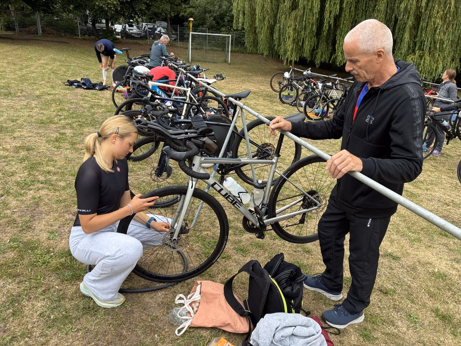 Junge Frau repariert Fahrradreifen, älterer Mann hält Fahrrad an Fahrradstall im Park.