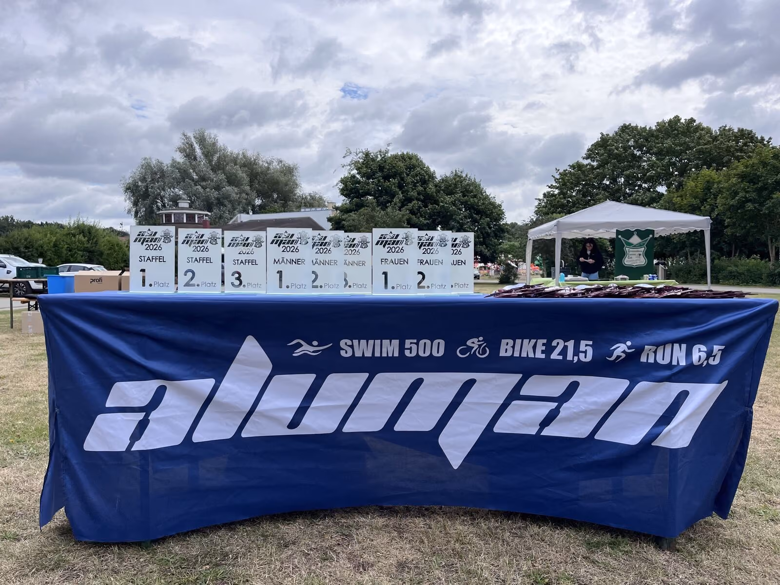 Tisch mit blauer Decke und Pokalen für Swim-Bike-Run-Wettbewerb Aluman 2026 im Freien.
