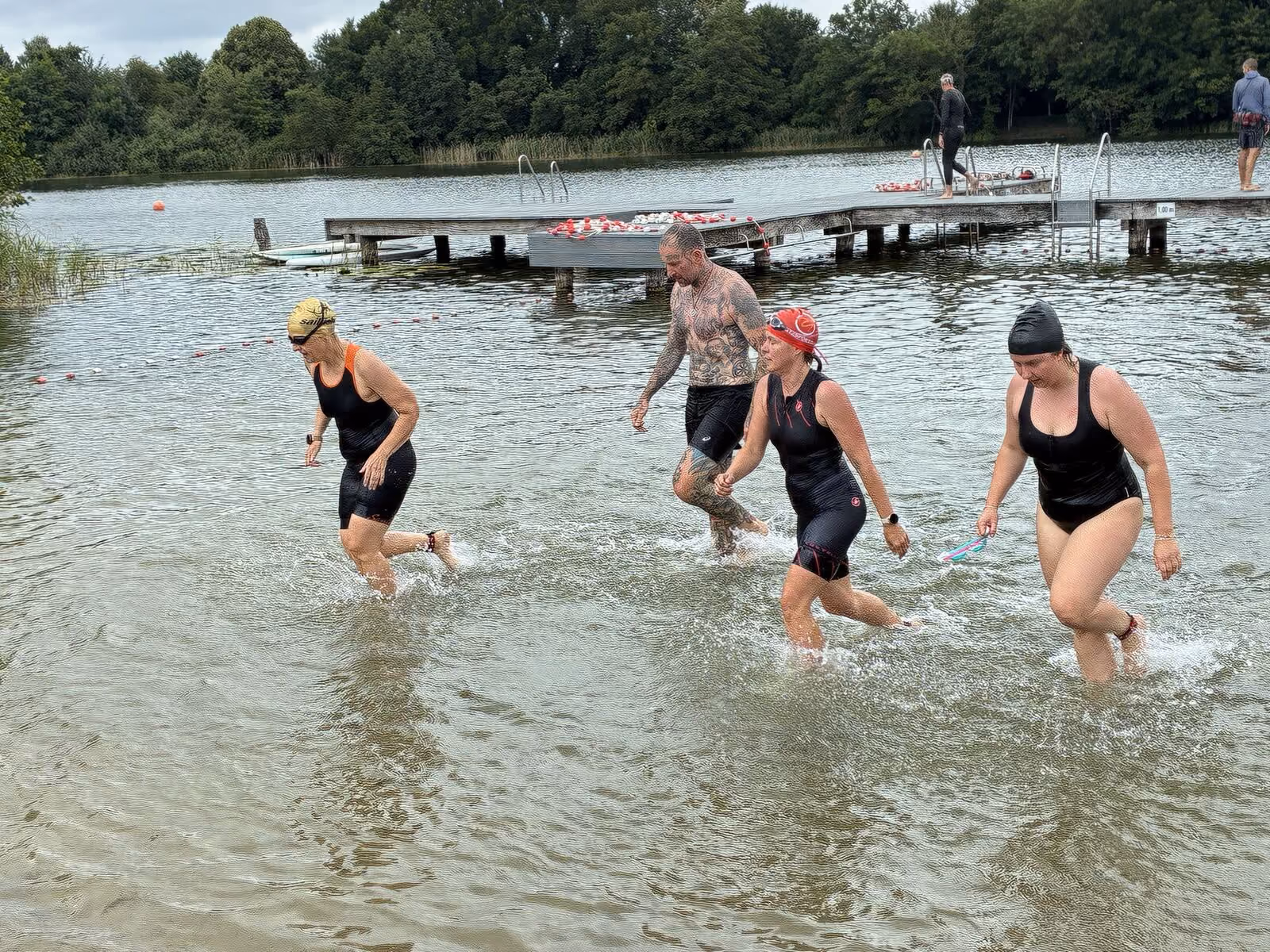 Vier Schwimmer mit Badekappen steigen aus einem See mit Steg und Bäumen im Hintergrund.