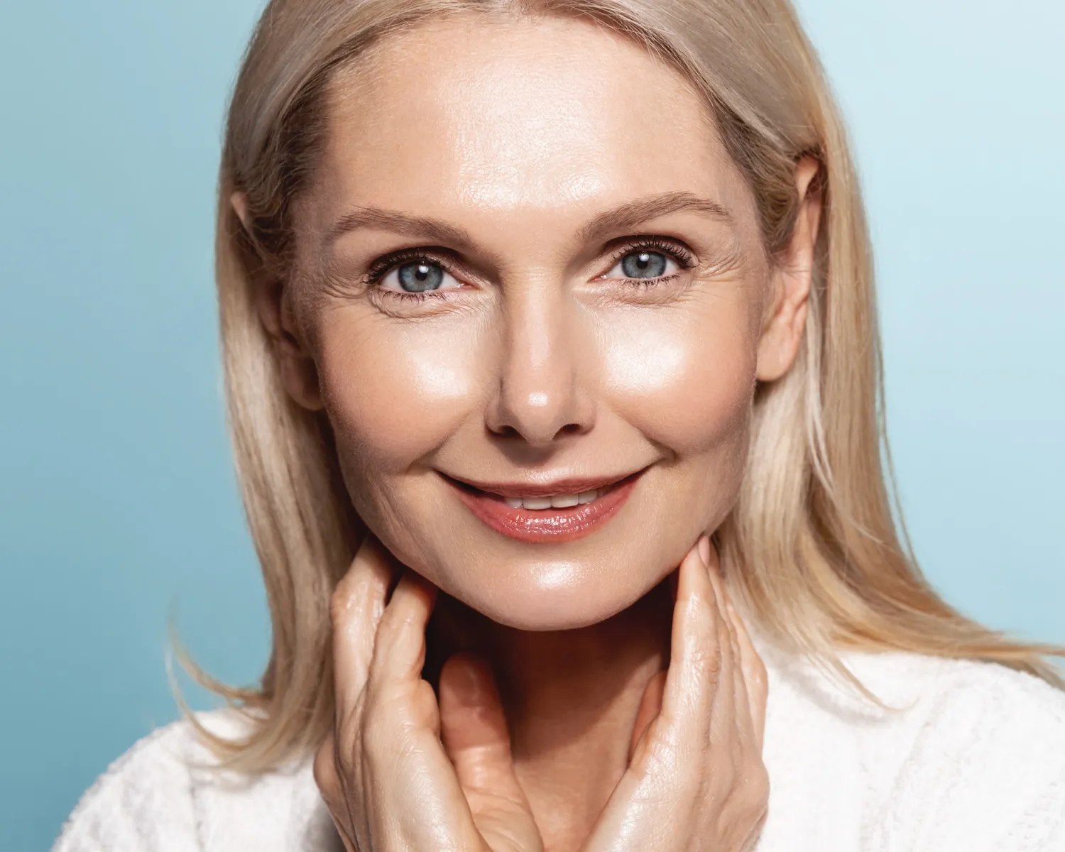 Close-up of a smiling mature woman with blonde hair and blue eyes touching her neck against a light blue background.