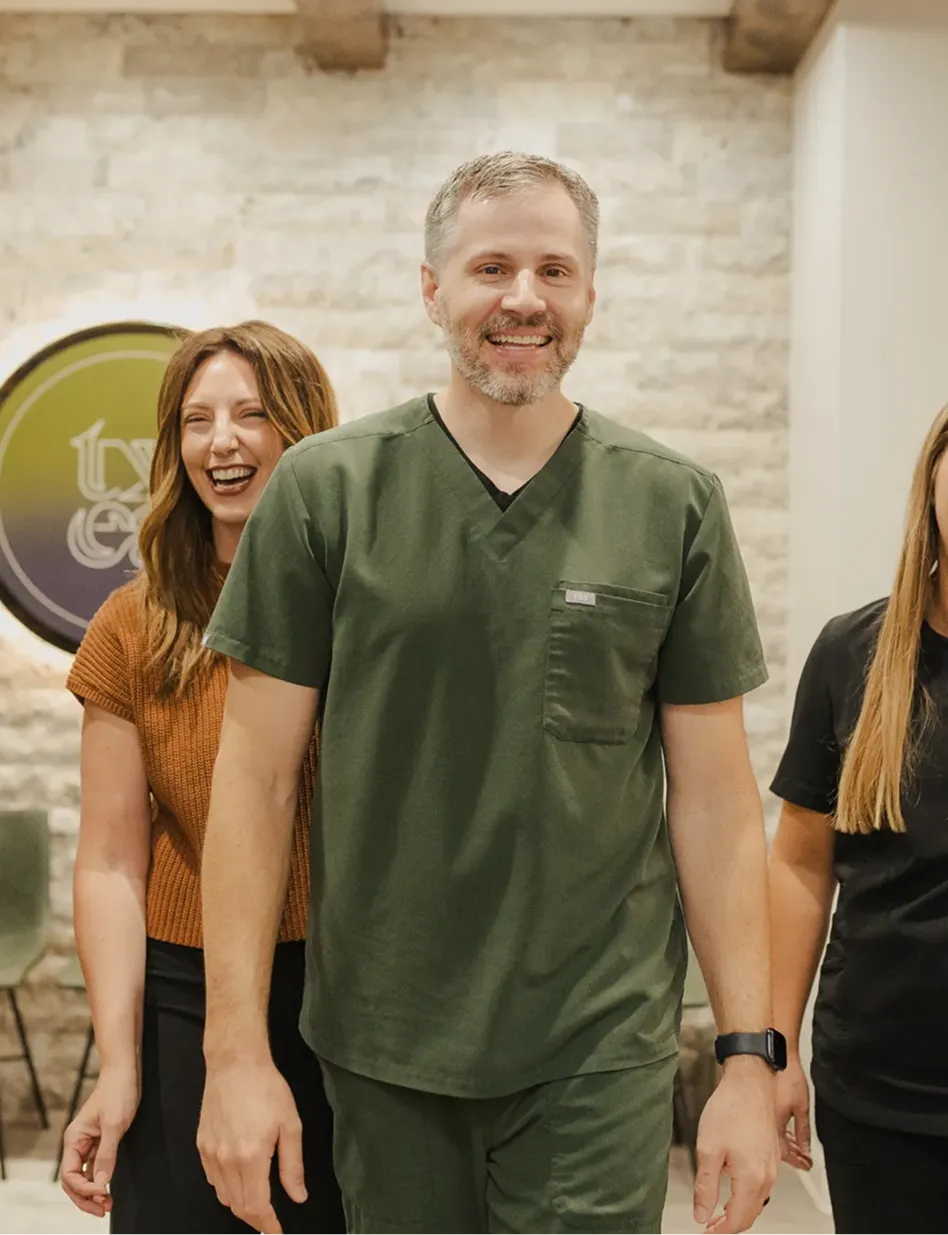 Smiling man in green scrubs walking indoors with two women, one laughing behind him.