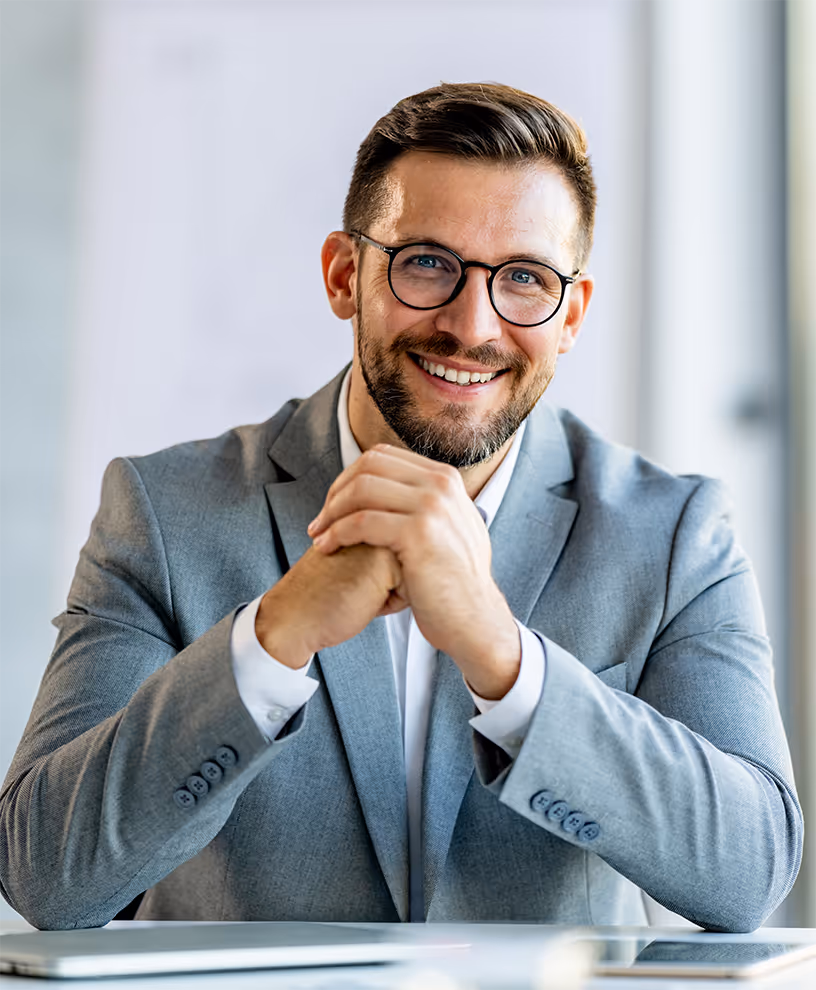 Smiling bearded aesthetic clinic professional wearing glasses, a light gray suit jacket, and white shirt, seated with hands clasped together on a table.