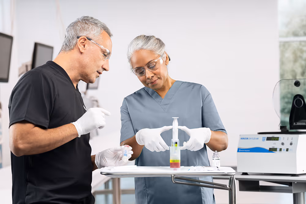 Two aesthetic medicine practitioners in scrubs and gloves work at a tray with a syringe of autologous blood for PRP treatment and a PRP centrifuge nearby.