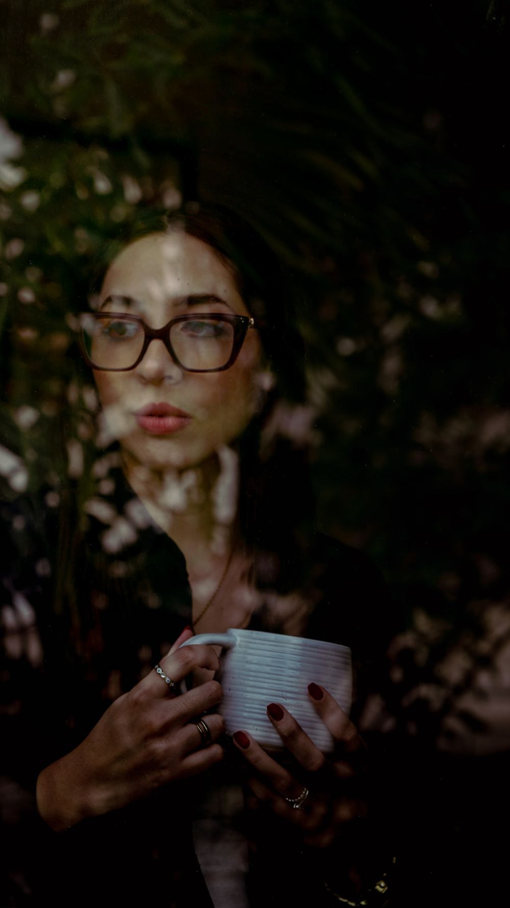 A woman with glasses holds a cup beside a window, seen through layered reflections.