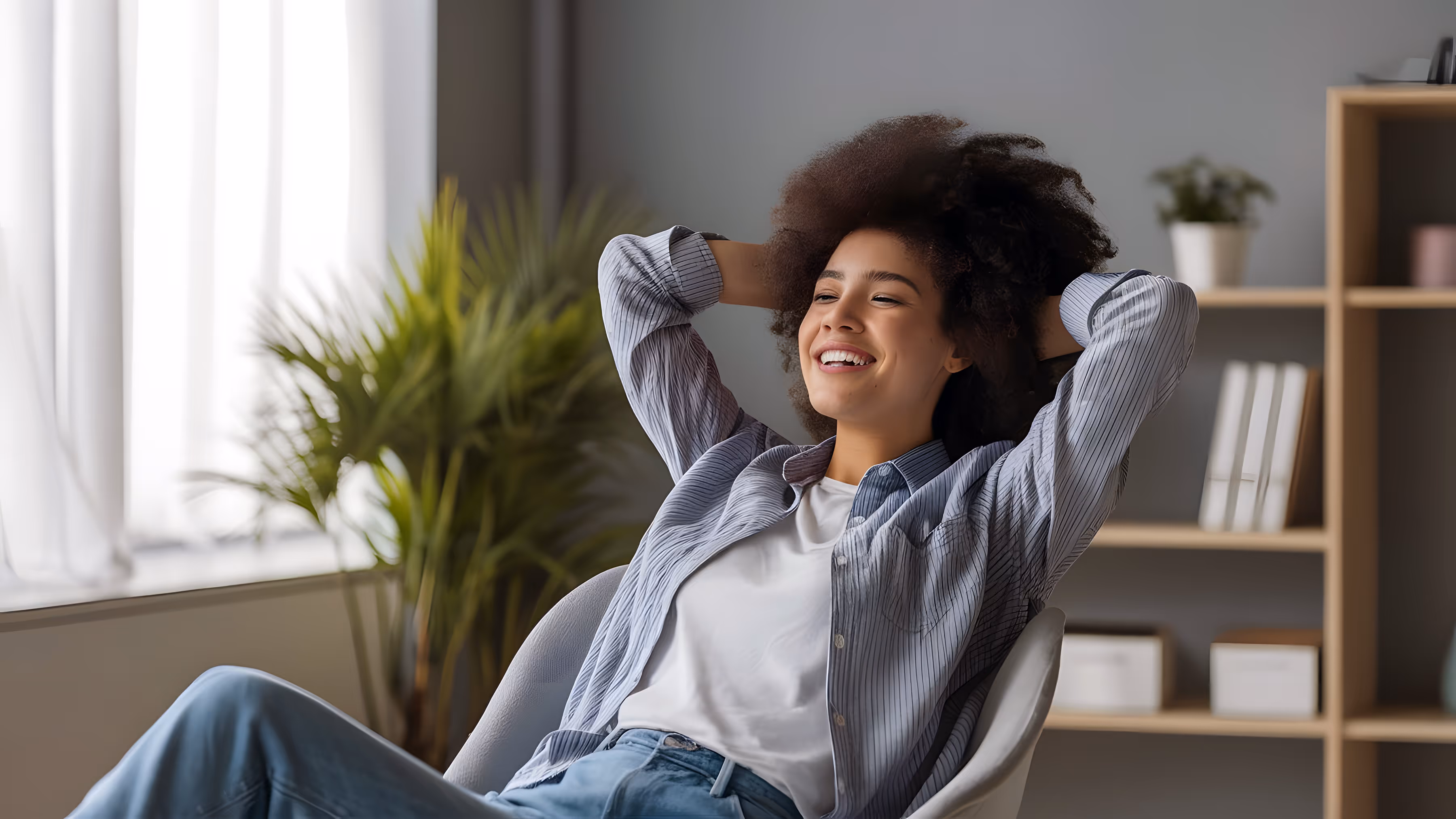 A woman in jeans, a blue sweater, and socks sitting on a yellow-cushioned chair with a laptop on her lap and stretching her arms.