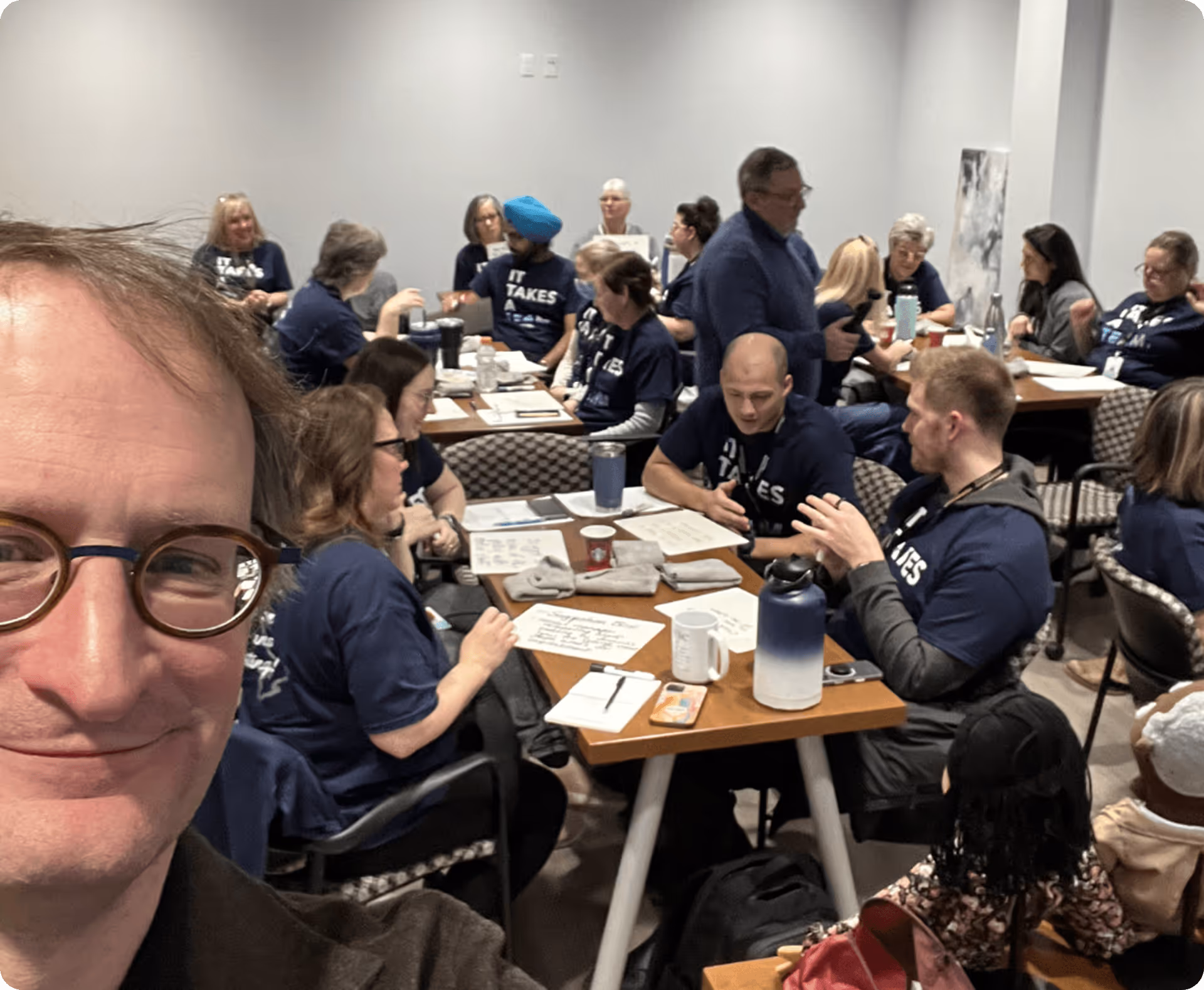 Group of people in a meeting room wearing matching navy shirts, talking and writing at tables.