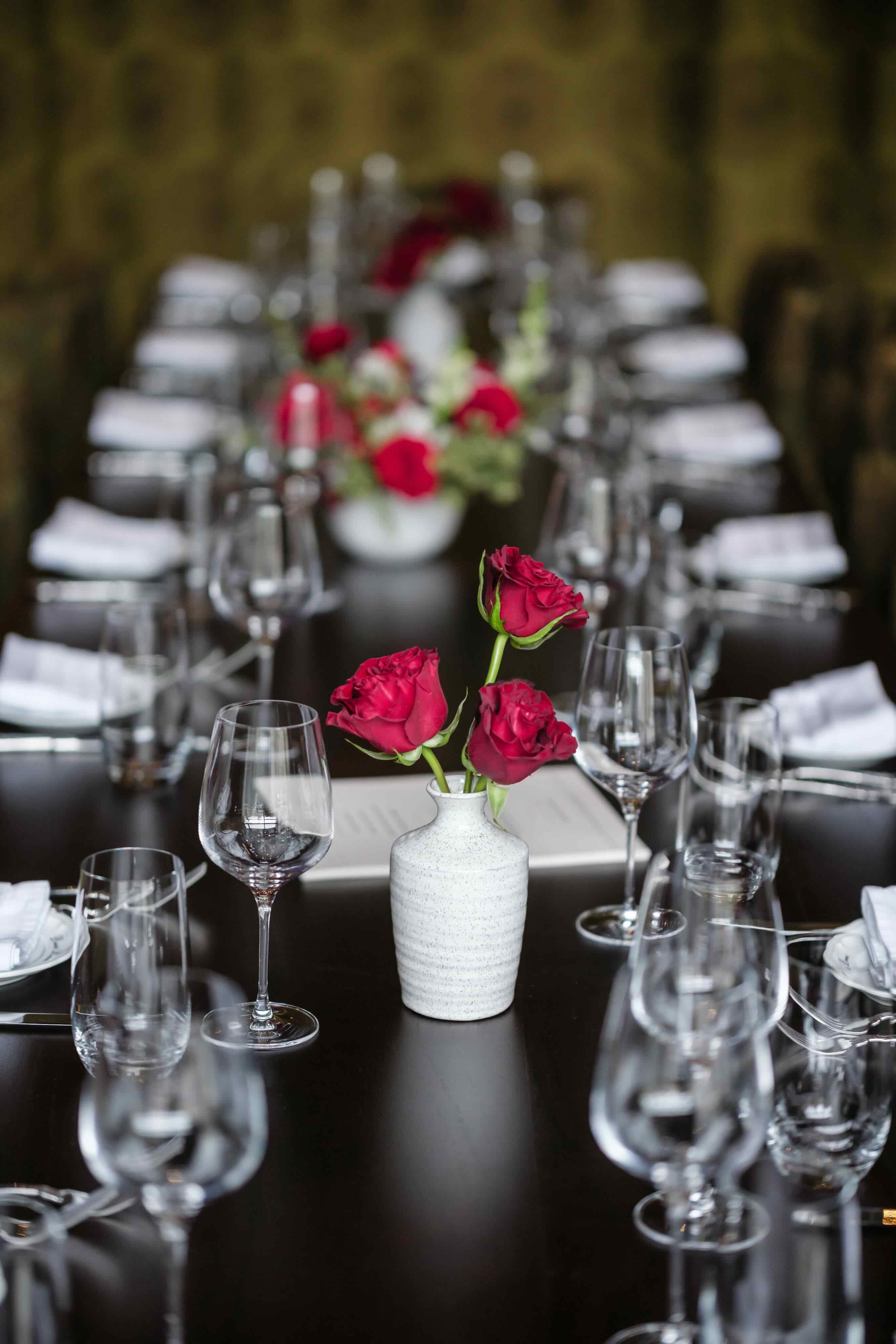 A long table with a vase of roses and wine glasses.