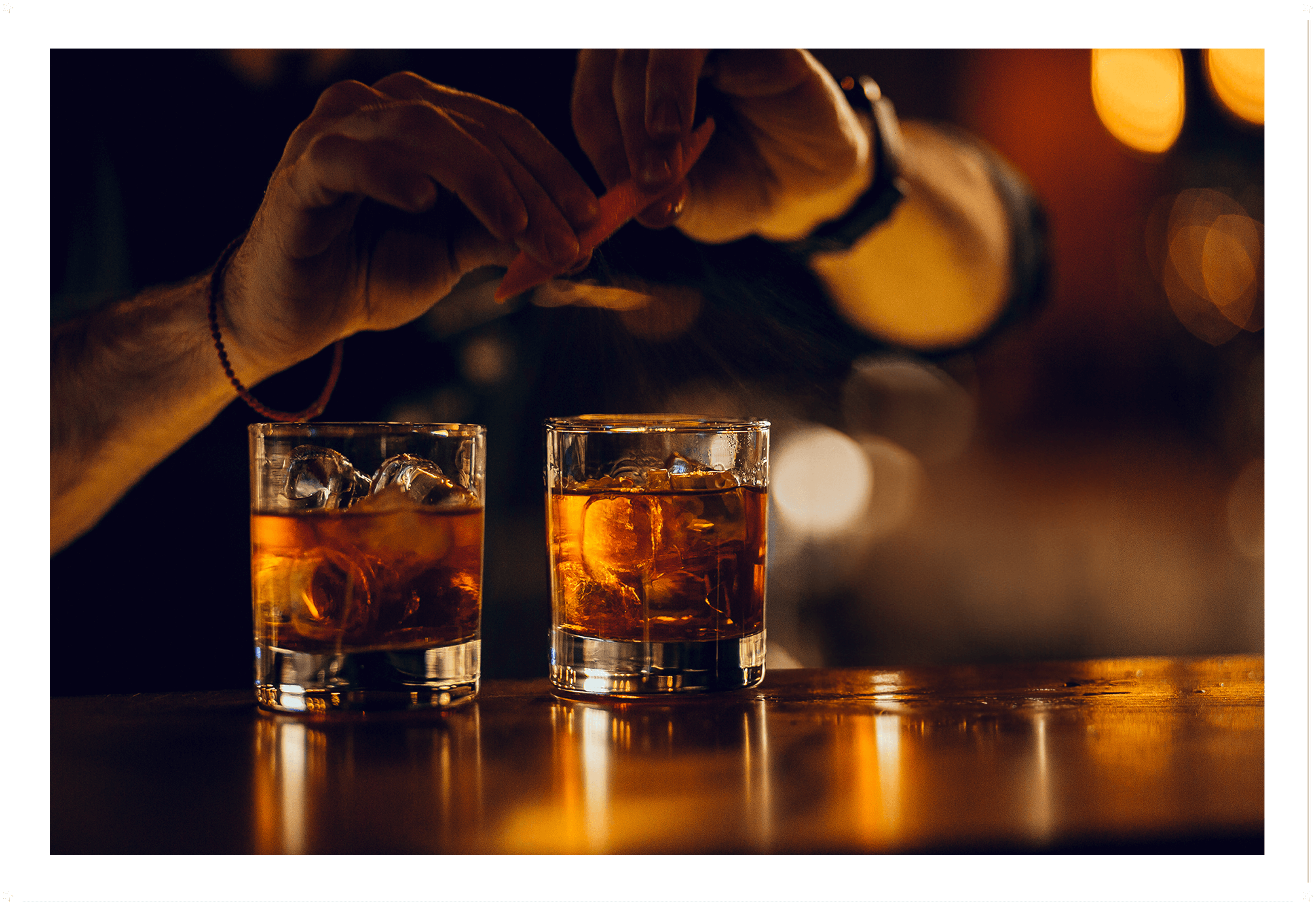 Close-up of two whiskey glasses with ice on a bar counter, one being garnished with an orange peel zest.