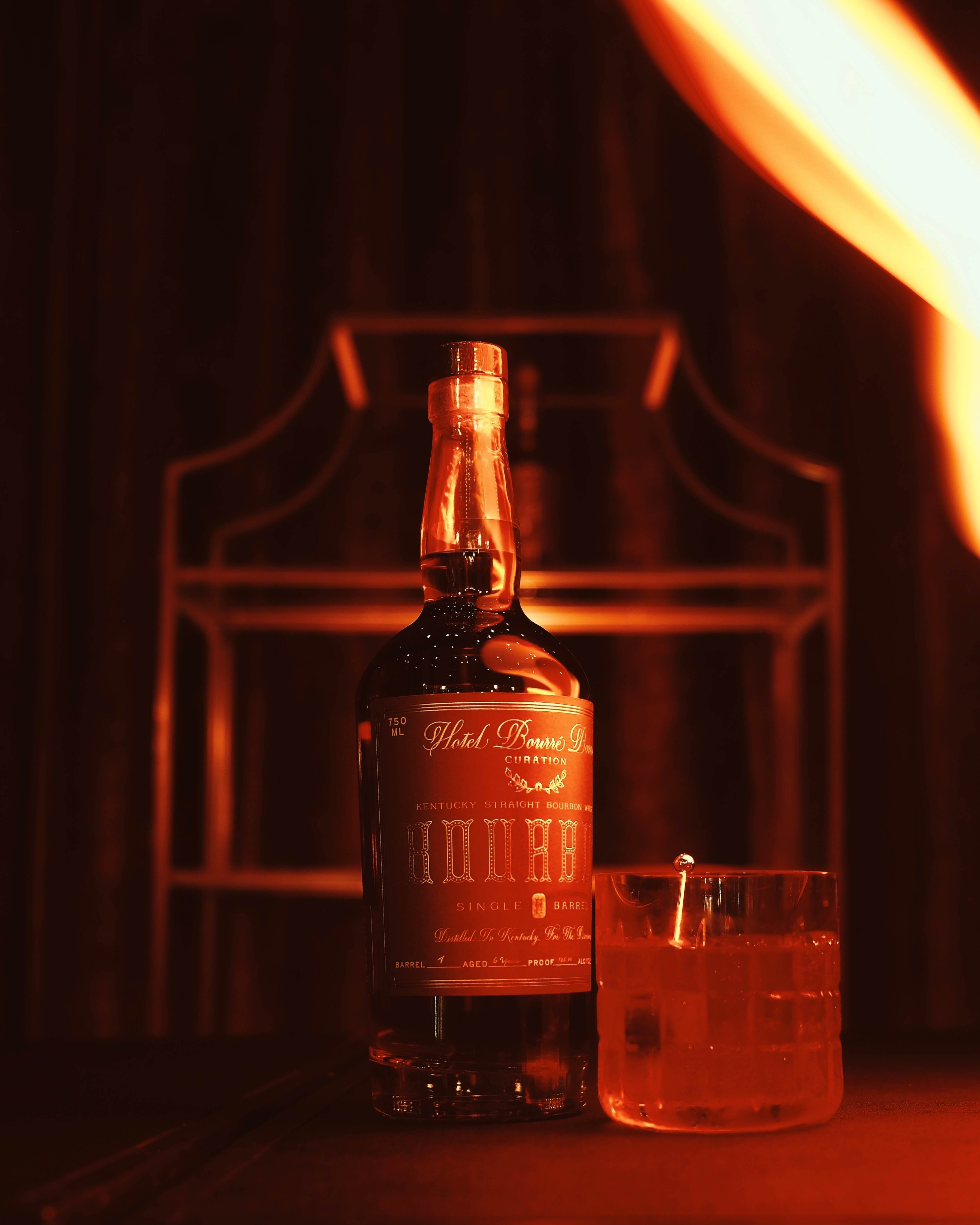 Bottle of Kentucky straight bourbon whiskey and a glass with ice on a dark surface, lit by warm, amber lighting.