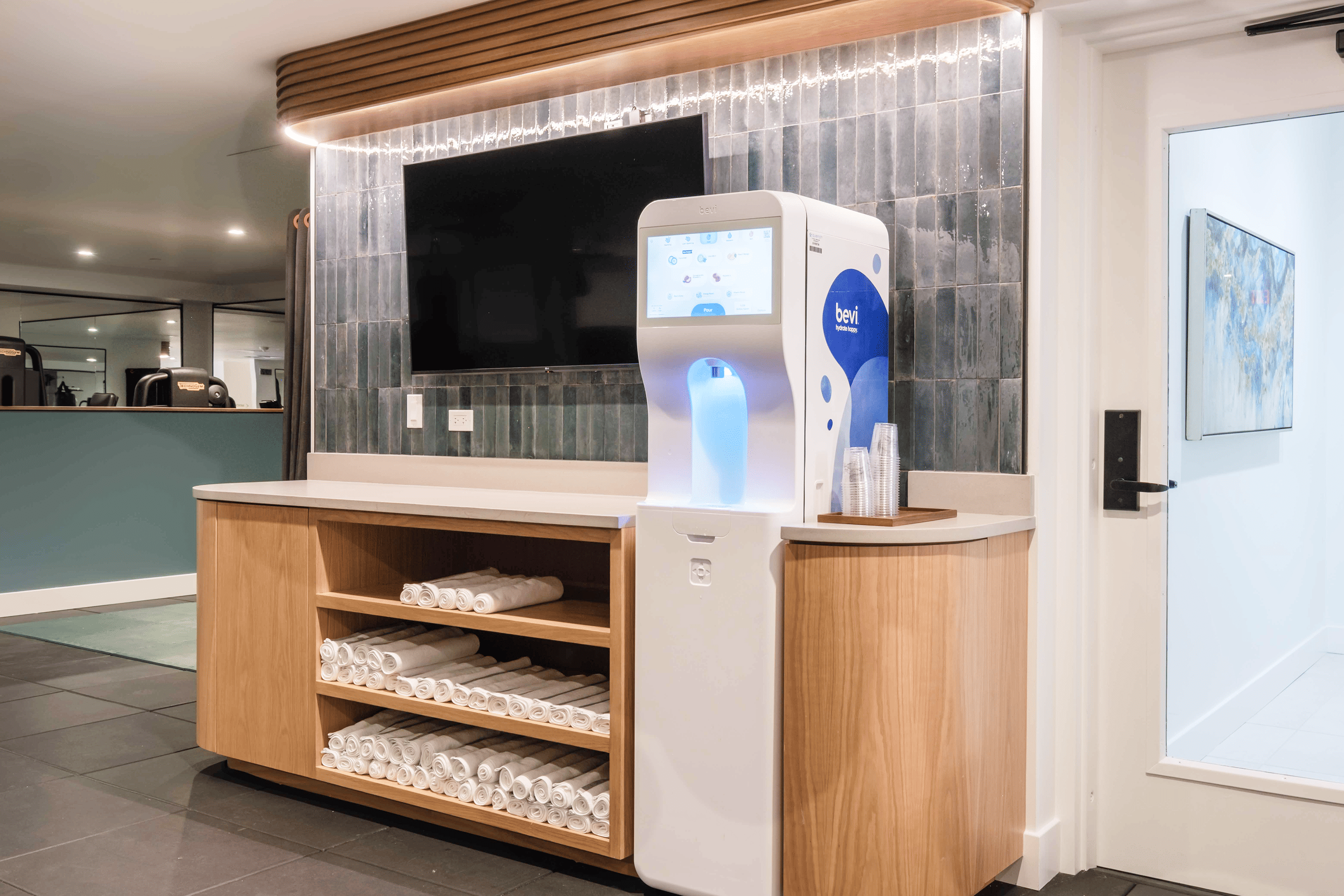 Modern hydration station with a touchscreen dispenser and stacked rolled towels on wooden shelves in a fitness center.