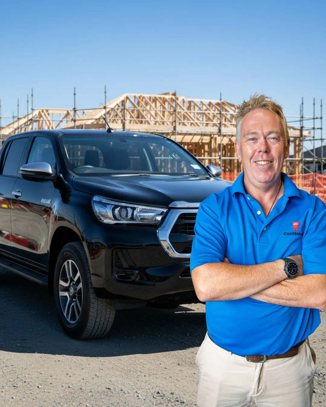 Man in a blue CarMoney polo standing with his arms crossed in front of a black Toyota Hilux, with a timber-framed house under construction behind him.