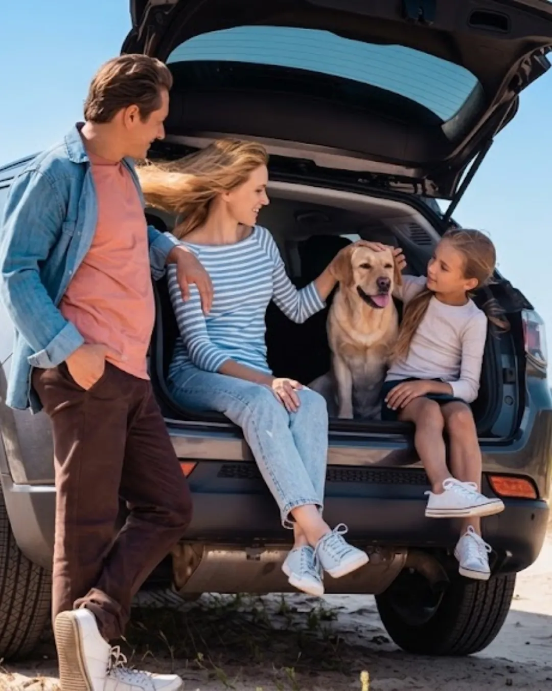 Family of three sitting and standing around the open boot of an SUV with a golden labrador, on a sunny day.