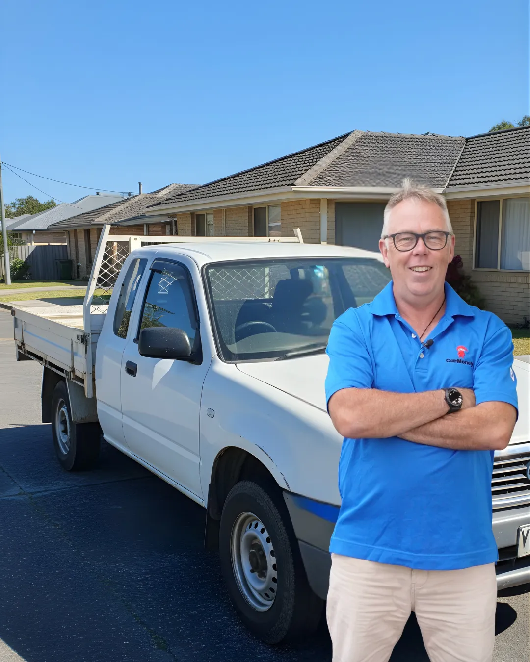 Man with glasses and a blue CarMoney polo standing with his arms crossed in front of a white flatdeck ute parked on a suburban street.