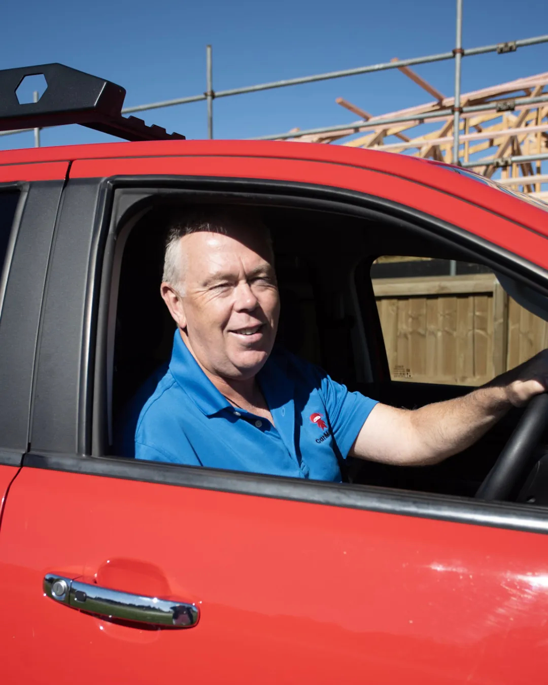 Smiling man in a blue CarMoney polo sitting in the driver’s seat of a red ute, with a timber-framed house under construction behind him.
