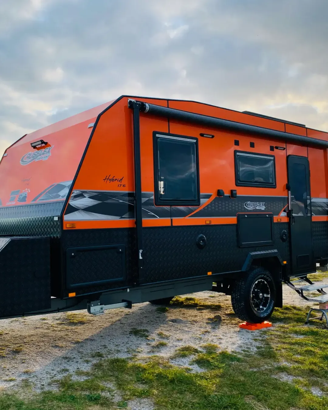 Orange and black Causeflex Hybrid off-road caravan parked on grass under a cloudy sky.