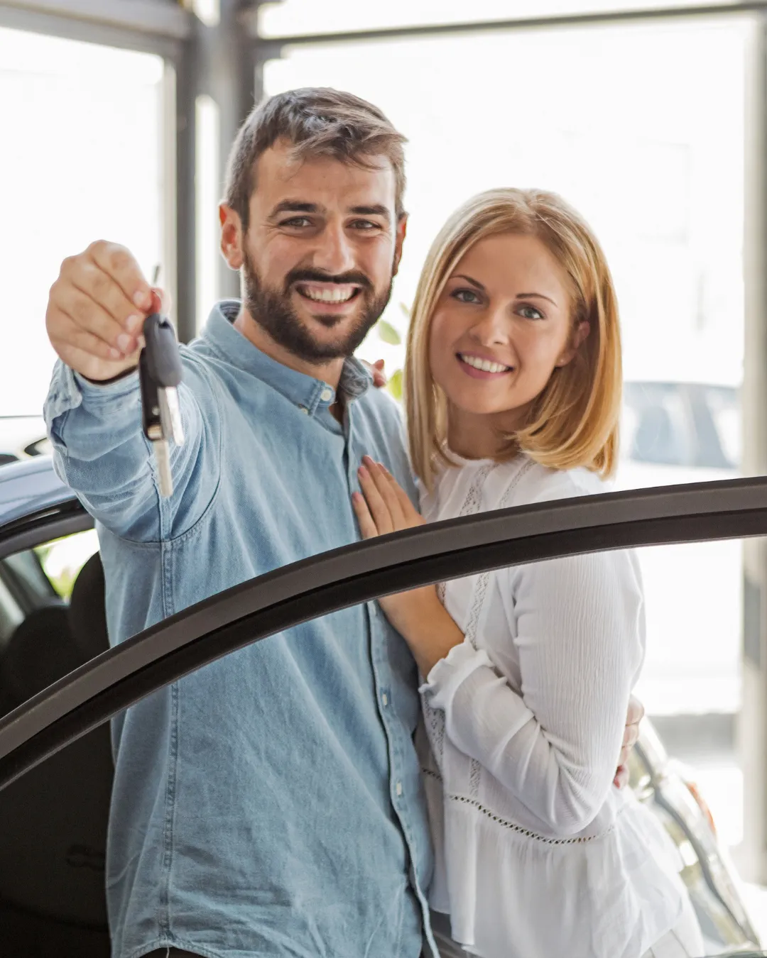 Smiling couple standing beside a car in a showroom, with the man holding up a set of car keys.