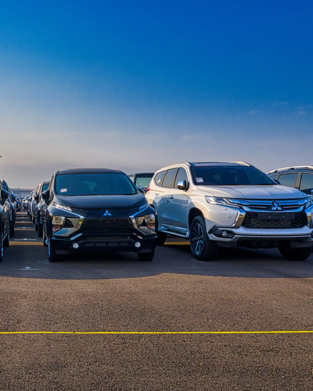 Rows of new Mitsubishi vehicles, including a black people mover and a silver SUV, parked in a lot under a clear blue sky.