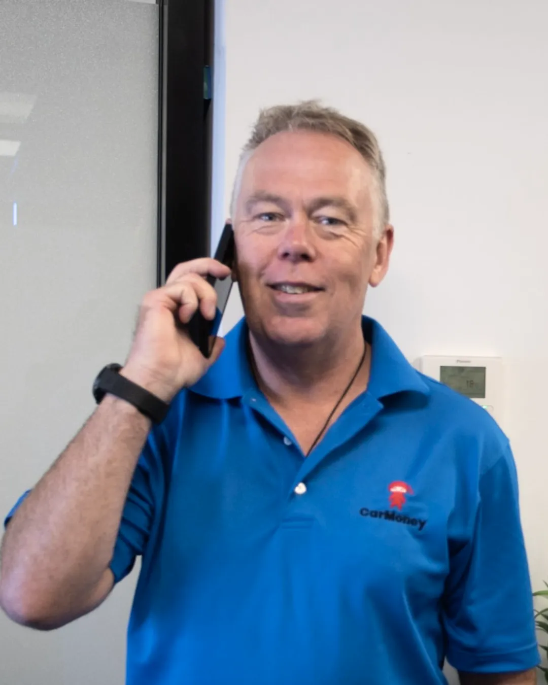 Man in a blue CarMoney polo smiling while talking on a mobile phone in an office.