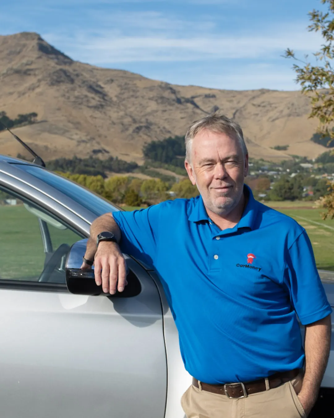 Man in a blue CarMoney polo leaning on the side mirror of a silver vehicle, with brown hills and green fields behind him.