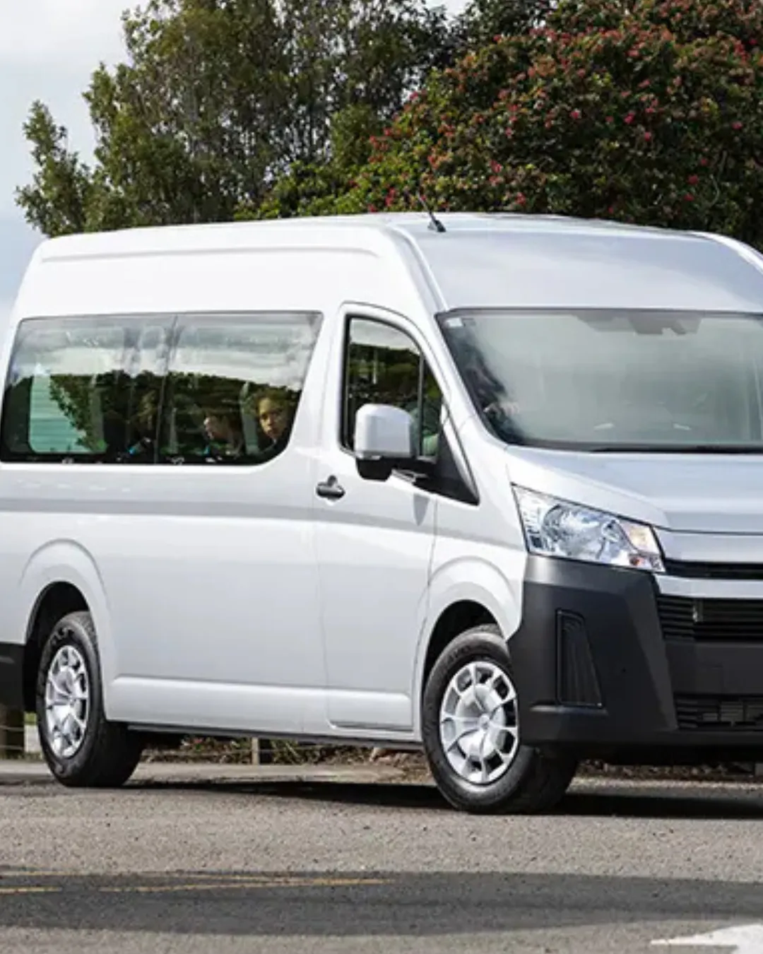 White Toyota HiAce minibus with passengers visible through the side window, parked on a road beside trees.